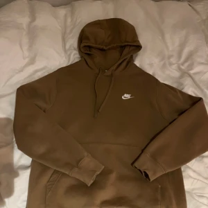 Brun hoodie från Nike - Säljer en snygg  hoodie från Nike med klassisk logga på bröstet. Tröjan har en bekväm passform och är perfekt för kyligare dagar. Den har en justerbar huva och en stor ficka framtill. Perfekt för en avslappnad stil.