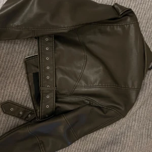 Oliv skinnjacka med bälte - Snygg svart skinnjacka med coola detaljer som dragkedjor och bälte i midjan. Perfekt för en edgy look. Jackan har långa ärmar och en klassisk krage.