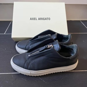 Svarta sneakers från Axel Arigato - Snygga svarta sneakers från Axel Arigato med dragkedja istället för traditionell snörning. Skorna har en stilren design med vit sula som kontrasterar mot det svarta skinnet. Perfekta för en modern och minimalistisk look. Storlek 42