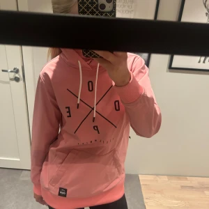 Rosa hoodie från Dope - Säljer en snygg rosa hoodie skidjacka från Dope med svart tryck på framsidan. Den har en stor ficka framtill och vita dragsnören vid huvan samt dragkedja vid armhålan för ventilation.