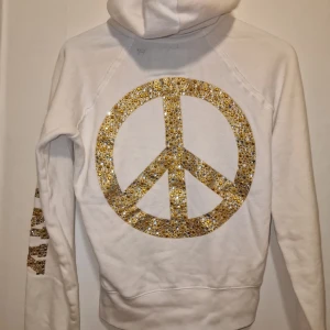Vit hoodie från Victoria's Secret  - Säljer en vit hoodie från Victoria's Secret med glittrande gulddetaljer. Den har en stor peace-symbol på baksidan och perfekt för att sticka ut. Finns inte att köpa längre! Garderobsrensning så kika gärna på övriga auktioner. 