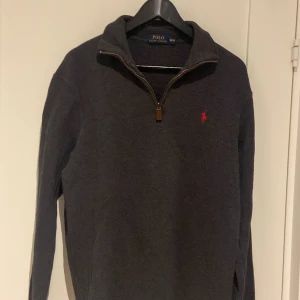 Mörkgrå tröja från Polo Ralph Lauren - Säljer en stilren mörkgrå tröja från Polo Ralph Lauren med en halv dragkedja och det klassiska röda logotypbroderiet på bröstet. Perfekt för en avslappnad men ändå snygg look. 