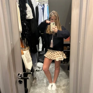 Beige prickig kortkjol - Säljer en söt beige kortkjol med bruna prickar och volangdetaljer. Kjolen har en lekfull design som passar perfekt för varmare dagar. Passar 36-38