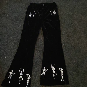 Svarta byxor med skelettmotiv - Unika svarta byxor med skelettmotiv i vitt. Byxorna har en loose passform och är dekorerade med skelettfigurer längs benen och benmotiv vid fickorna. Perfekta för en djärv och lekfull stil.