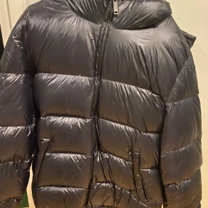 Svart pufferjacka från Zara - Säljer en svart pufferjacka från Zara med dragkedja och huva. Jackan är perfekt för kyliga dagar och har en glansig yta som ger en modern look. Den är designad för att hålla dig varm och bekväm.