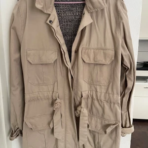 Beige parkajacka med knytband - Säljer en stilren beige parkajacka med knytband i midjan och flera praktiska fickor framtill. Jackan har långa ärmar och en stickad insida i grått. Perfekt för höst och vår och även sommar!