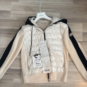 Moncler Padded Tricot Cardigan With Hood In White And Navy Blue - - Köpt på Nk i april 2024. Kvitto och alla andra tillbehör fås med. Använd 2-3 gånger (som ny). Nypris 12499kr. Cardiganen är väldigt limiterad och är väldigt svår att få tag på. Senaste bud 5500kr. OBS! Byte är bara i intressse om kläderna är i nyskick och kvitto finns. Söker efter en vanlig svart Moncler cardigan (utan luva, svart logga).