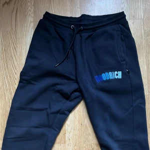 Svarta mjukisbyxor från Hoodrich - Snygga svarta mjukisbyxor från Hoodrich med broderad logga i blå nyanser. Byxorna har snörning i midjan och praktiska fickor på sidorna. Perfekta för en avslappnad stil.