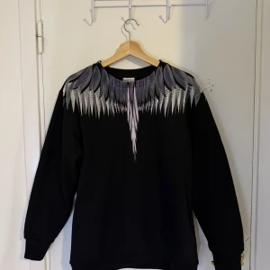 Marcelo Burlon Tröja - Säljer en svart tröja från Marcelo Burlon med ett unikt fjädermönster i grått och vitt runt halsen. Tröjan har långa ärmar och en avslappnad passform. Perfekt för dig som vill ha något stilrent men med en twist.