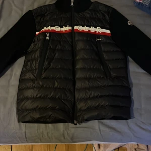 Svart dunjacka från Moncler - Snygg svart cardigan från Moncler med broderad logga på ärmen och textdetalj över bröstet. Cardiganen har en dragkedja framtill och två praktiska dragkedjor till fickorna.