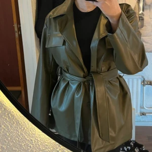 Olivgrön skinnjacka med bälte - Snygg olivgrön skinnjacka med knytbälte i midjan. Jackan har långa ärmar och en stilren design med knappar framtill. Perfekt för att ge en cool touch till din outfit.