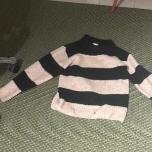 Randig stickad tröja - Säljer en mysig stickad tröja med breda ränder i svart och beige. Tröjan har en normal passform och långa ärmar, perfekt för kyligare dagar. Från Gina i storlek 158-164
