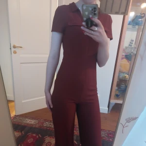 Tegelröd 70's jumpsuit från ASOS - Säljer en snygg brun jumpsuit från ASOS med korta ärmar och krage. Jumpsuiten har en dragkedja framtill och är i ett ribbat material som ger en skön passform. Perfekt till den som letar efter en 70tals-look!