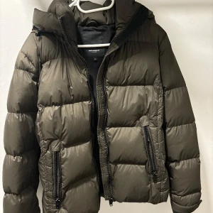 Olivgrön dunjacka från Rockandblue - Säljer en olivgrön dunjacka från Rockandblue. Jackan har en pufferdesign med dragkedja och huva, perfekt för kyliga dagar. Den har två framfickor med dragkedjor och en diskret logga på baksidan. En stilren och funktionell jacka för vintern. Nypris 3600kr.