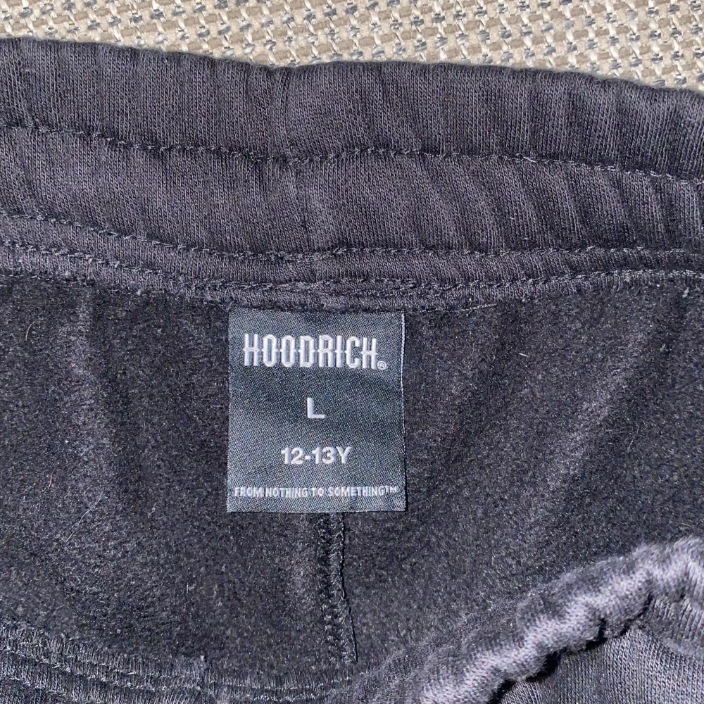 Säljer ett par svarta mjukisbyxor från Hoodrich med elastisk midja och snörning. Byxorna har en avslappnad passform och logotypen 'Hoodrich' tryckt i vitt och blått på sidan. Perfekta för en bekväm stil.. Farkut & Housut.