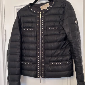 Svart lättviktsjacka med nitar - Snygg svart dunjacka från Michael kors med coola nitar längs dragkedjan och fickorna. Jackan har en rund halsringning och långa ärmar. Perfekt för att ge din outfit en edgy touch.