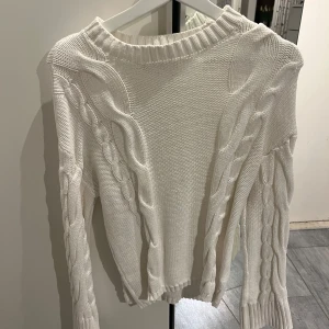 Vit stickad tröja med flätmönster - Säljer en stilren vit stickad tröja med snyggt flätmönster längs ärmarna. Tröjan har långa ärmar och ribbade kanter vid hals. Från Lager 157 