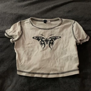 Vit croppad topp med fjärilstryck från Divided - Säljer en vit croppad topp från Divided med ett coolt fjärilstryck i svart på framsidan. Toppen har korta ärmar och kontrastsömmar i svart. Perfekt för en trendig look!