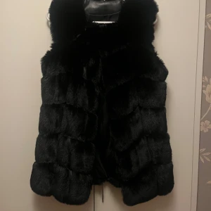 Svart pälsväst - Snygg svart pälsväst som ger en lyxig känsla. Perfekt för att hålla sig varm och stilren under kallare dagar. Västen har en mjuk och fluffig textur som gör den extra mysig. Köpt för 699:-. Passar storlek 34/36.