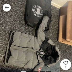 Grå dunjacka från Canada Goose - Säljer en grå dunjacka från Canada Goose med broderad logotyp på ärmen. Jackan har en justerbar huva och dragkedja framtill. Perfekt för kalla dagar! 🧥❄️