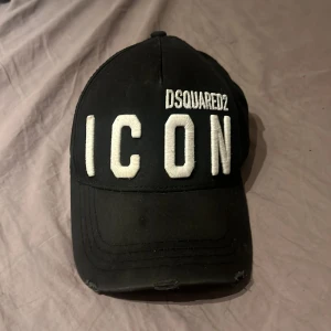 Svart keps från Dsquared2 - Snygg svart keps från Dsquared2 med stor vit broderad text 'ICON' på framsidan. Justerbar rem baktill för perfekt passform. Nypris ligger på cirka 1700kr men jag säljer den för 800kr men priset går att diskutera!