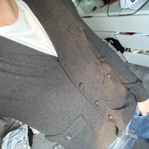 Mörkgrå cardigan - Säljer en stilren mörkgrå cardigan med knappar framtill. Cardiganen har långa ärmar och praktiska fickor på båda sidor.