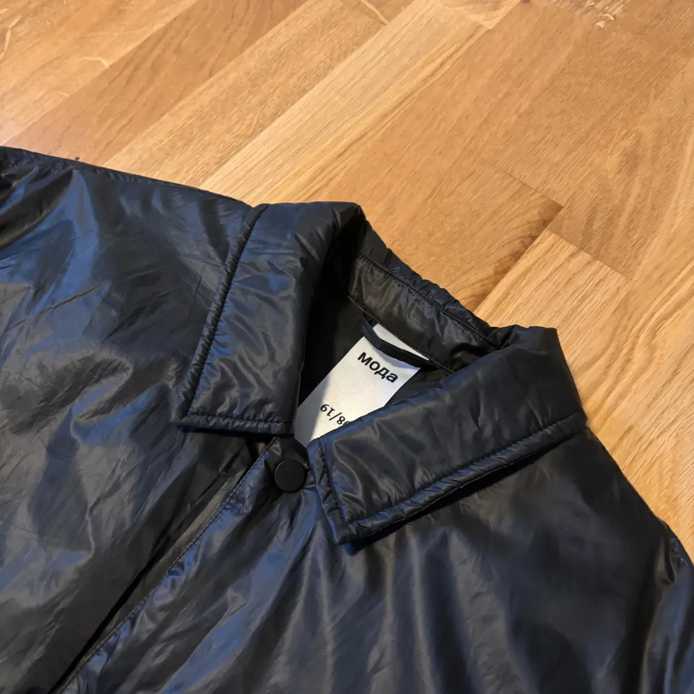 Säljer en stilren svart fodrad vindjacka från MADEBYMODA . Jackan har en klassisk krage och knäppning framtill. Den är utrustad med en bröstficka och har en diskret broderad detalj på bröstet . Takit.