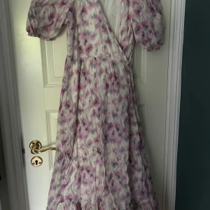 Blommig omlottklänning från & Other Stories - Säljer en lång, blommig omlottklänning från & Other Stories med puffärmar och volang nertill. Klänningen har lila och rosa toner på vit botten och knyts i midjan. 