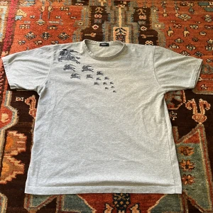 Grå Burberry t-shirt - Säljer en grå t-shirt från Burberry med asfett mörkblått tryck på. T-shirten har rund hals och är kortärmad. En perfekt oversized och stilren tröja🌟