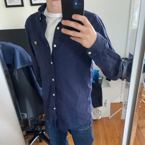 Mörkblå skjorta från Lyle & Scott - Säljer en mörkblå skjorta från Lyle & Scott med klassisk passform. Skjortan har långa ärmar, knappar framtill och en liten gul broderad logga på bröstet. Perfekt till jeans eller chinos för en clean look.