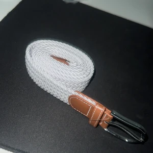 Vitt flätat bälte med bruna skinn-detaljer - Snyggt vitt flätat bälte med bruna skinn-detaljer och silverfärgat spänne. Perfekt accessoar för att lyfta din outfit med en stilren touch. Bältet har klassisk design och passar till många olika looks.