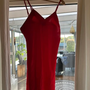 Röd klänning med smala axelband från Jolynn - Säljer en röd klänning från Jolynn med smala axelband och v-ringad halsringning. Klänningen är tillverkad i 100% polyester och har en enkel, elegant design som passar perfekt för festliga tillfällen.