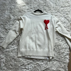 Vit stickad tröja med rött hjärta från Ami - Säljer en stilren vit stickad tröja från Ami med ett rött hjärta och bokstaven A broderat på bröstet. Tröjan har rund halsringning och ribbade muddar vid ärmslut och nederkant. Perfekt för dig som gillar klassisk design med en twist.