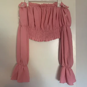 Rosa offshoulder blus från Amisu - Säljer en söt rosa offshoulder blus från Amisu med långa vida ärmar och smockad midja. Blusen har volangdetaljer vid ärmslut och axlar, perfekt för en trendig look.