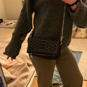 Svart axelväska med nitar och kedja - Säljer en svart axelväska i skinnimitation med silverfärgade nitar på locket och en snygg kedjerem. Väskan har dragkedja och två spännen framtill. Perfekt för att lyfta din outfit med lite edge. Från even&odd och är i bra skick! 💕