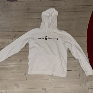 Vit hoodie från Sail Racing - Säljer en vit hoodie från Sail Racing med svart logga tryckt på bröstet. Tröjan har huva och en stor magficka framtill. Klassisk och enkel design, perfekt för dig som gillar stilrena plagg. Den är i fint skick använd endast några få gånger och inte urtvättad priset kan justeras vara skriv om du har några frågor!