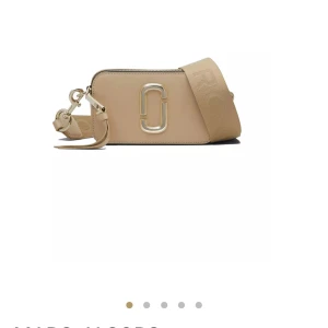 Beige axelväska från Marc Jacobs - Snygg beige liknande Marc Jacobs väska med bred justerbar axelrem och ikonisk logga i metall framtill. Väskan har dragkedja upptill och detaljer i guld. Perfekt för att bära det viktigaste med stil.