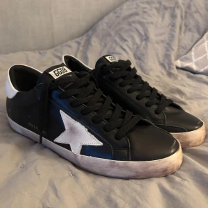 Svarta sneakers från Golden Goose med vit stjärna - Säljer ett par svarta Golden Goose sneakers med vit stjärndetalj på sidan och vit häl. Skorna har svarta skosnören, GGDB-logga på plösen och en något smutsad sula för den klassiska looken. Skorna har lite slitage inget som stör! Hör av dig om du har några frågor! 