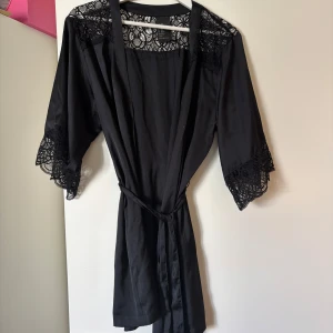 Svart kimono med spetsdetaljer från HM - Säljer en svart kimono från HM med vackra spetsdetaljer vid axlar och ärmslut. Kimonon har trekvartsärm och knyts i midjan med ett bälte. Perfekt att slänga på sig hemma eller till mysiga kvällar.
