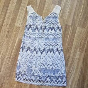 Mönstrad sommarklänning i blått och vitt - Säljer en ärmlös kortklänning med zigzag-mönster i blå och vita toner. Klänningen har v-ringning med knytning fram och är tillverkad i ett lätt syntetmaterial, perfekt för varma dagar.