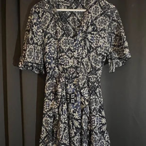 Blåvit mönstrad klänning från Zara - Säljer en blåvit klänning från Zara med kort ärm och vackert blommigt och geometriskt mönster. Klänningen har v-ringning och markerad midja för en smickrande passform. Perfekt för vår och sommar!