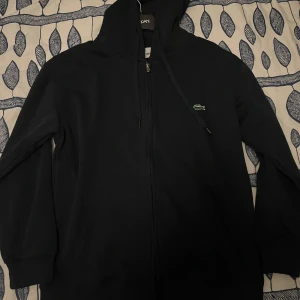 Svart hoodie med dragkedja från Lacoste - Säljer en svart hoodie från Lacoste med dragkedja och huva. Klassisk krokodil-logga på bröstet och justerbara snören i huvan. Regular fit och långärmad modell, perfekt för en avslappnad stil.