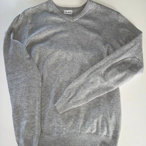 Grå stickad tröja med v-ringning från John henric  - Säljer en klassisk grå stickad tröja från john henric med v-ringad hals. Tröjan har långa ärmar och ribbade muddar vid ärmslut och nederkant. Perfekt för lager-på-lager och enkel att matcha med det mesta.