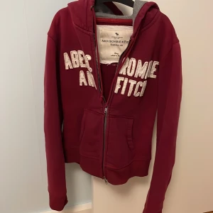 💓Vinröd Abercrombie & Fitch hoodie💓 - Mysig vinröd hoodie från Abercrombie & Fitch med vit broderad logga framtill. Jackan har dragkedja, två fickor och grått innerfoder i luvan. Perfekt för en avslappnad stil och enkel att matcha med jeans eller leggings.💓