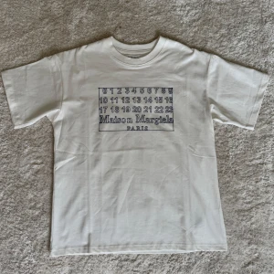Maison Margiela T-shirt - Vit t-shirt från Maison Margiela med svart tryck framtill som visar siffror och logotypen. Klassisk rund hals och normal passform. Perfekt för dig som gillar stilrena och trendiga plagg. Passar M-L