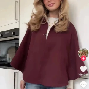 Vinröd capejacka med knapp - Säljer en vinröd capejacka med vid passform och en knapp framtill. Jackan har en lös och oversized siluett, perfekt att slänga över en stickad tröja. Passar dig som gillar en stilren och trendig look.