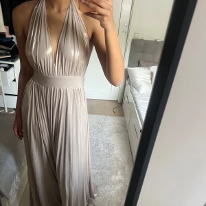 Champagnefärgad långklänning med halterneck - Säljer en glansig champagnefärgad långklänning med halterneck och djup v-ringning. Klänningen har öppen rygg, markerad midja och plisserad kjol. Stängs med dragkedja i sidan. Perfekt till balen💕 nypris:1600kr 