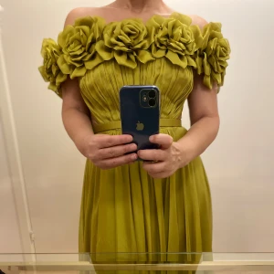 Grön långklänning med stora blommor - Säljer en maxilång grön klänning med offshoulder-modell och stora 3D-blommor längs överdelen. Klänningen har markerad midja med bälte och plisserad kjol som faller snyggt. Perfekt för bal eller fest.