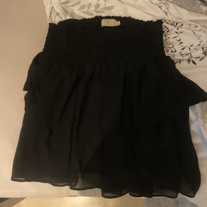 Svart kortkjol från By Chelsea - Svart kortkjol från By Chelsea med smockad midja och lätt, luftig känsla. Kjolen har en volangig nederdel som ger ett flowy intryck. Perfekt för dig som gillar en enkel men stilren look. Köpte för ett tag sen men knappt använd 