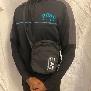 Svart axelväska från Emporio Armani EA7(oanvänd), vi kan diskutera pris - Snygg svart axelväska från Emporio Armani EA7 med vitt tryck på framsidan. Väskan har justerbar axelrem, dragkedja och en praktisk ytterficka och 3 innerfickor. Perfekt för att bära med sig det viktigaste.